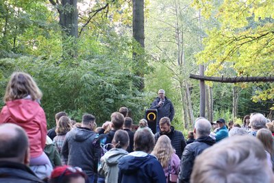 Eine große Menschengruppe steht vor einem Mann, der in ein Mikro spricht